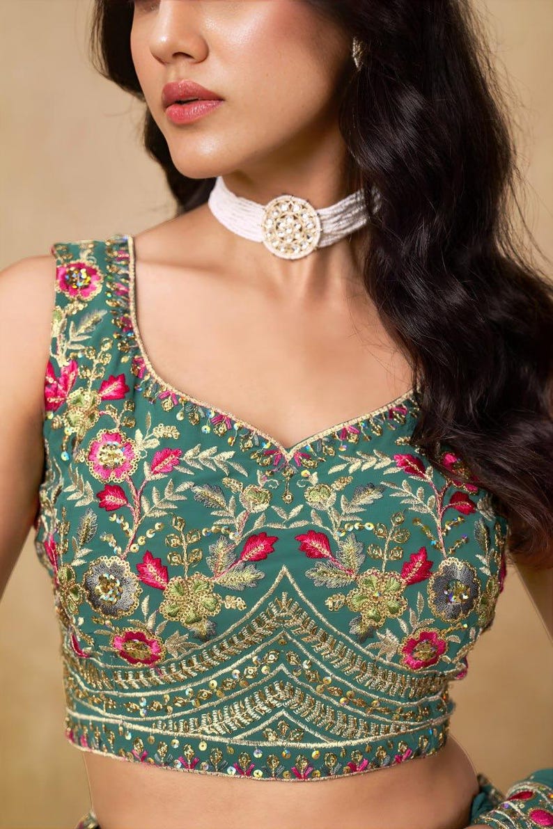 May include: Emerald green sleeveless top with intricate floral embroidery in pink, gold, and green. The top features a scoop neckline and is adorned with sequins. A white beaded choker necklace with a gold pendant completes the look. This is a traditional Indian garment.