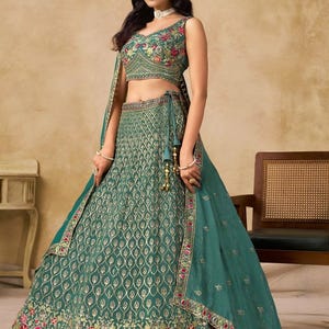 May include: A teal-colored, sleeveless crop top and skirt set with intricate gold embroidery and floral accents. The skirt features a detailed pattern and a decorative border. A matching teal scarf is draped over the shoulder. The outfit is suitable for special occasions.