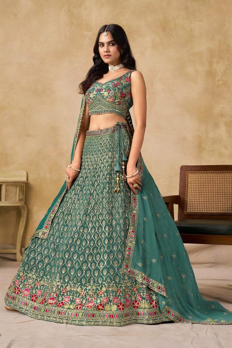 May include: A teal-colored, sleeveless crop top and skirt set with intricate gold embroidery and floral accents. The skirt features a detailed pattern and a decorative border. A matching teal scarf is draped over the shoulder. The outfit is suitable for special occasions.