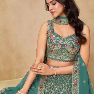May include: Teal-colored embroidered formal dress with a matching dupatta. The sleeveless top features floral embroidery in pink and gold. The skirt has a detailed pattern with gold accents. The outfit is accessorized with gold jewelry.