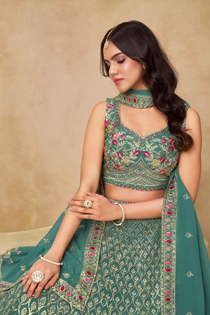 May include: Teal-colored embroidered formal dress with a matching dupatta. The sleeveless top features floral embroidery in pink and gold. The skirt has a detailed pattern with gold accents. The outfit is accessorized with gold jewelry.