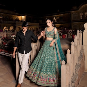 Puede incluir: Un conjunto de lehenga color verde azulado con una blusa sin mangas y una falda larga con intrincados bordados dorados y detalles florales rosas, junto con una dupatta transparente a juego. Una camisa negra y pantalones claros.