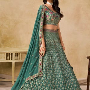 May include: Emerald green lehenga with intricate gold embroidery and floral accents. The ensemble includes a matching dupatta and a cropped top with floral embroidery. The skirt features a detailed pattern and a decorative border.