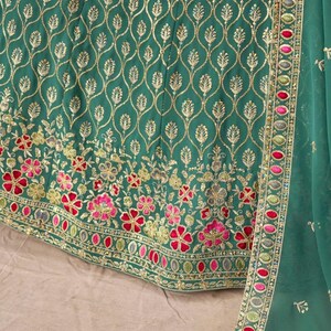 May include: Close-up of a teal green garment with intricate gold embroidery. The design features a repeating pattern of gold leaves and floral embroidery in pink, red, and gold. A matching teal green fabric with a decorative border is visible.