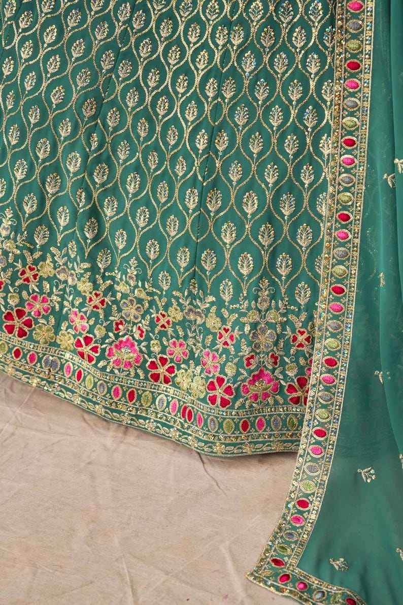 May include: Close-up of a teal green garment with intricate gold embroidery. The design features a repeating pattern of gold leaves and floral embroidery in pink, red, and gold. A matching teal green fabric with a decorative border is visible.