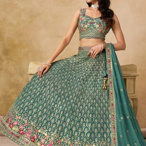 May include: A teal green lehenga choli set with intricate gold embroidery. The sleeveless top and flared skirt feature a repeating pattern, with floral detailing in pink and red at the hem. A matching dupatta completes the ensemble.