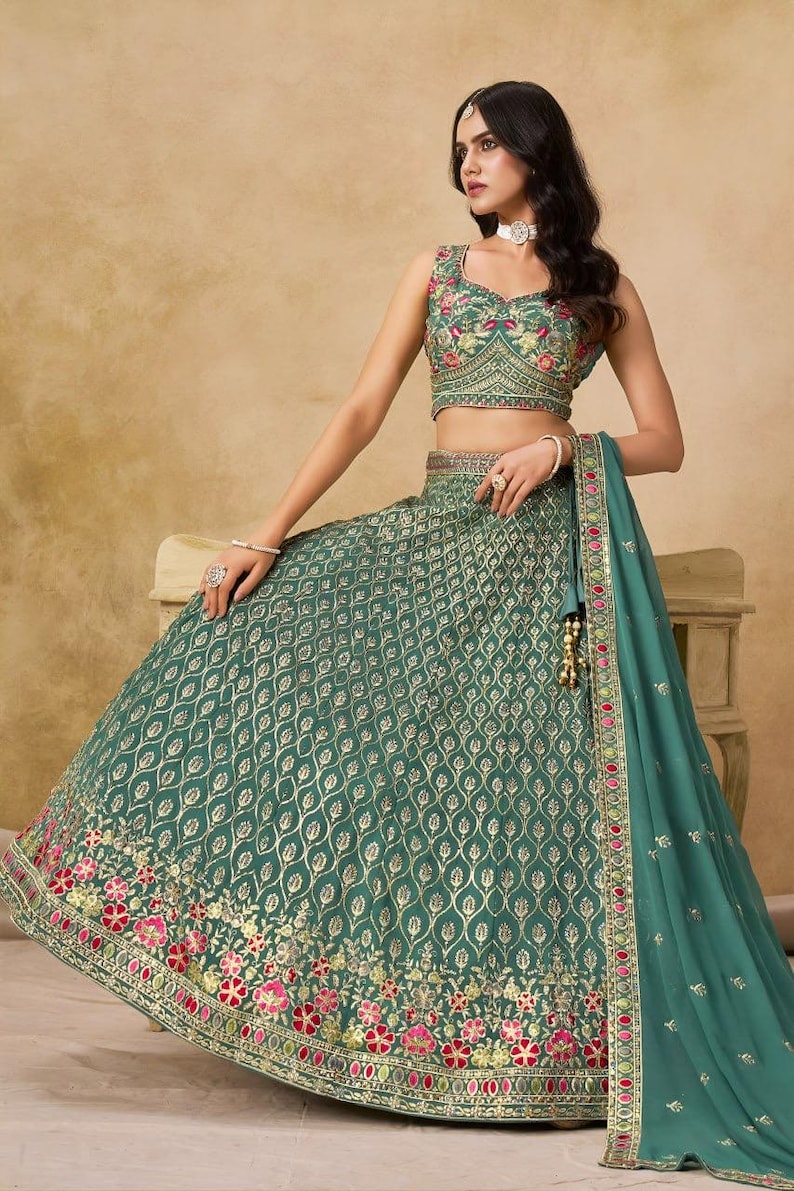May include: A teal green lehenga choli set with intricate gold embroidery. The sleeveless top and flared skirt feature a repeating pattern, with floral detailing in pink and red at the hem. A matching dupatta completes the ensemble.