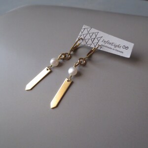 May include: Gold earrings with a small white pearl on each earring. The earrings have a long, thin, gold bar hanging from the pearl. The earrings are on a white card with the text "InfinEight handmade in Canada".