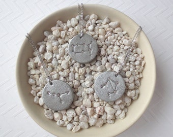 Può includere: Tre collane d'argento con ciondoli a forma di costellazione zodiacale su sfondo bianco. I ciondoli sono a forma di cerchio con le costellazioni dei Gemelli, della Bilancia e del Cancro.