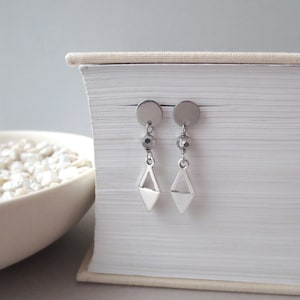 May include: A pair of silver earrings with a geometric diamond shape dangle and a small silver bead. The earrings are displayed on a white book.