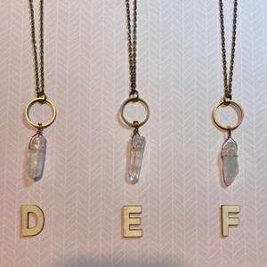 May include: Three necklaces with clear quartz crystal pendants. Each pendant is wire-wrapped and hangs from a gold-colored ring and chain. The background is a pink and white chevron pattern. Wooden letters D, E, and F are at the bottom.