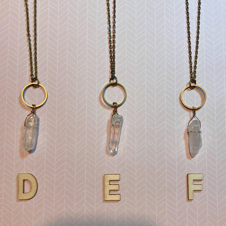 May include: Three necklaces with clear quartz crystal pendants. Each pendant is wire-wrapped and hangs from a gold-colored ring and chain. The background is a pink and white chevron pattern. Wooden letters D, E, and F are at the bottom.