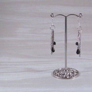 Op de afbeelding: Een paar zilveren oorhangers met zwarte kralen die aan een ketting hangen. De oorhangers worden getoond op een zilveren standaard.