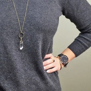 May include: A gray V-neck sweater is paired with blue jeans. A gold-colored necklace with a clear crystal pendant hangs over the sweater. A black watch and gold jewelry adorn the wrist.