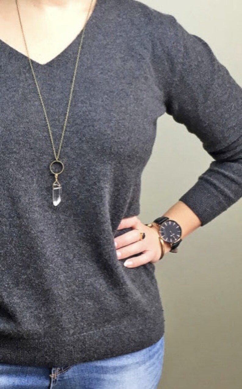 May include: A gray V-neck sweater is paired with blue jeans. A gold-colored necklace with a clear crystal pendant hangs over the sweater. A black watch and gold jewelry adorn the wrist.