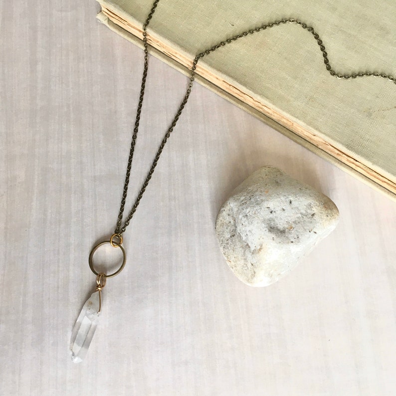 May include: A long necklace with a clear quartz crystal pendant, a gold-colored ring, and a dark chain. A small, light-colored stone and a book are also in the image. The necklace is a piece of jewelry.