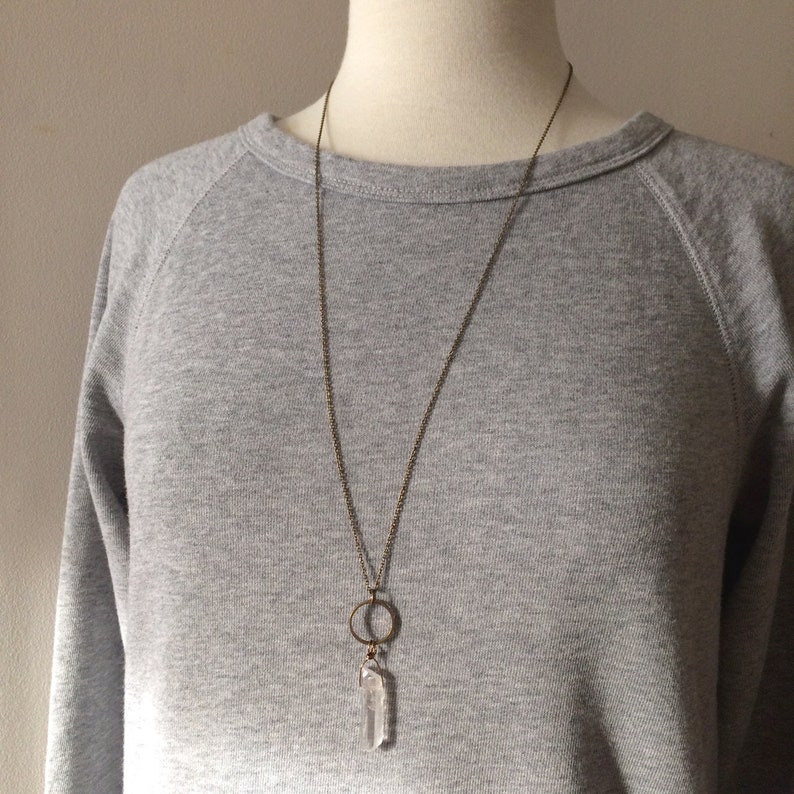 May include: A long, antique brass necklace featuring a clear quartz crystal pendant. The pendant hangs from a delicate chain and includes a small brass circle. The necklace is displayed on a mannequin wearing a gray sweatshirt.
