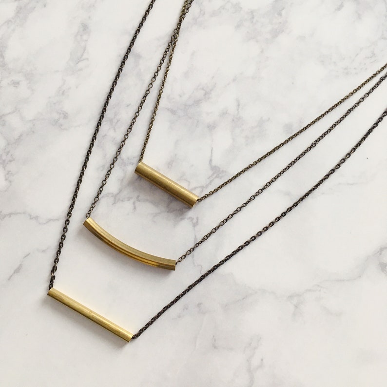Puede incluir: Un collar de oro de varias capas con tres estilos de cadena diferentes y tres formas de colgantes geom&eacute;tricas. Las formas de los colgantes son una barra curva, una barra recta y un cilindro corto.