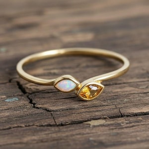 May include: A gold ring featuring two marquise-cut gemstones, one white and one yellow. The ring's band curves gently, and the gemstones are set close together. The ring is displayed on a rustic wooden surface.