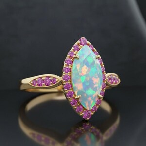 Può includere: Un anello d'oro con un'opale a taglio marquise, circondata da piccole pietre rosa. La fascia dell'anello presenta un design delicato con pietre rosa aggiuntive su entrambi i lati della pietra principale. L'anello è ambientato su uno sfondo scuro.