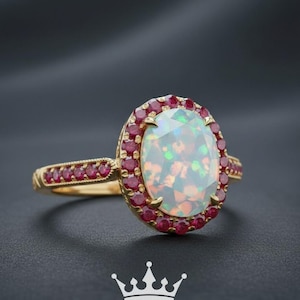 May include: A gold ring featuring an oval opal center stone surrounded by small red gemstones. The ring's band is also adorned with red gemstones. The ring is set against a dark background. The text "Sundaram Jewelers" is at the bottom.