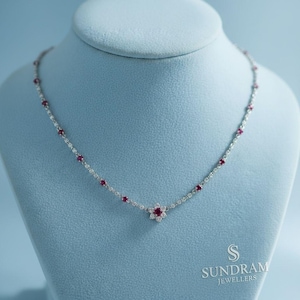 May include: A delicate silver necklace with a floral pendant and small red gemstones. The necklace is displayed on a blue mannequin. The necklace has a flower pendant with a cluster of red gemstones in the center.
