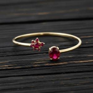 Könnte beinhalten: Ein goldfarbenes, offenes Armband mit zwei roten Edelsteinen. Einer ist oval, der andere ein Cluster kleinerer Steine in Sternform. Das Armband liegt auf einer dunklen Holzoberfläche.