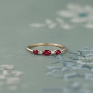 Op de afbeelding: Een delicate gouden ring met drie ovaal geslepen, dieprode edelstenen, geflankeerd door twee kleinere, ronde, heldere edelstenen. De ring heeft een dunne, gepolijste band, tegen een zachte, bloemige achtergrond.