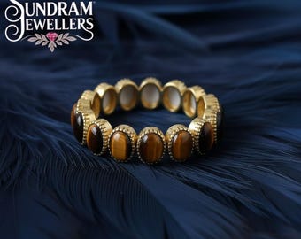 May include: A gold-colored ring with oval tiger's eye stones and white stones. The ring is set against a dark blue background. The Sundram Jewellers logo is in the upper left corner.
