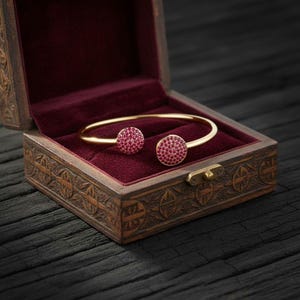 Puede incluir: Una pulsera dorada con dos adornos circulares de color rubí, presentada en un joyero de madera tallada abierto, forrado con terciopelo rojo. La caja se cierra con un pequeño cierre dorado.