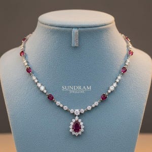 Può includere: Una collana d'argento con pendente e dettagli in rubini e diamanti. La collana presenta un pendente a goccia di rubino circondato da diamanti. La collana è esposta su un manichino blu. Il marchio "SUNDRAM JEWELLERS" è visibile.