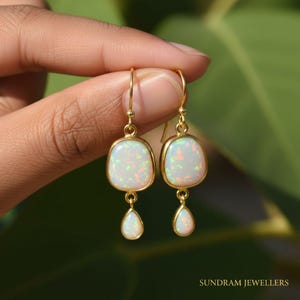 May include: Gold-tone earrings with white opal stones. Each earring features a rectangular top stone and a teardrop-shaped dangle. The opals have a milky white base with flashes of color. The earrings are held against a blurred green background.