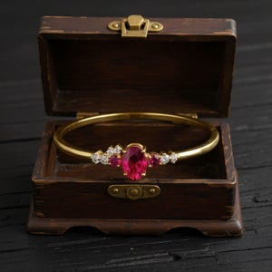 Può includere: Un bracciale rigido color oro con una grande pietra centrale ovale di rubino e piccoli accenti di rubino e diamanti. Il bracciale è esposto all'interno di un piccolo portagioie antico in legno aperto con chiusura in ottone.