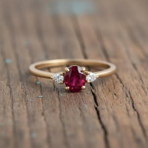 Puede incluir: Un anillo de oro con una gran piedra central de rubí ovalada y dos diamantes en forma de pera a cada lado. El anillo está sobre una superficie de madera, mostrando el diseño y el color de la joya.