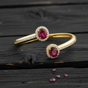 Puede incluir: Una pulsera de puño abierta de color dorado con dos piedras preciosas ovaladas de color rojo intenso, rodeadas por un halo de pequeñas piedras transparentes. La pulsera tiene un diseño delicado con pequeñas piedras transparentes a lo largo de la banda. Varias piedras preciosas sueltas están dispersas debajo.