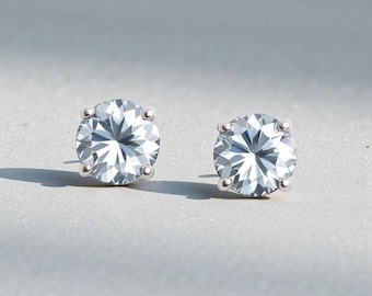May include: A pair of round, clear stud earrings. Each earring features a large, faceted, clear gemstone set in a silver-colored metal. The gemstones have a brilliant cut, reflecting light. The earrings are displayed on a light gray surface.