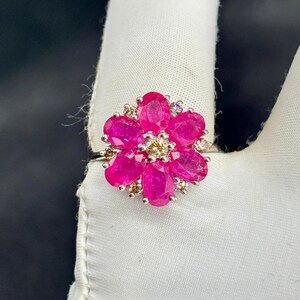 Puede incluir: Un anillo de plata con un diseño floral. La flor está compuesta por piedras preciosas rosas brillantes de talla ovalada, con piedras más pequeñas, claras y color champán que acentúan los pétalos. El anillo está sobre un fondo blanco.
