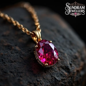 May include: A gold necklace featuring a ruby-red, oval gemstone pendant. The pendant is set in a gold bezel and suspended from a fine gold chain. The background is a dark, textured surface. The logo "Sundram Jewellers" is visible.