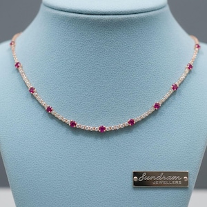 Op de afbeelding: Een roodgouden ketting met rode en heldere edelstenen. De ketting wordt gepresenteerd op een blauwe achtergrond. De edelstenen zijn klein en rond. Een klein rechthoekig plaatje met de tekst "Sundram Jewellers" is zichtbaar.