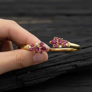 Puede incluir: Un brazalete abierto de color dorado con un grupo de piedras preciosas ovaladas de color rojo intenso y pequeños diamantes blancos. El brazalete se muestra sobre un fondo oscuro y texturizado, resaltando el diseño y el color de la joya.