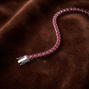 Op de afbeelding: Een tennisarmband met een doorlopende rij ronde, dieprode edelstenen gezet in een zilverkleurige metalen band. De armband is voorzien van een veilige baksluiting aan één uiteinde en heeft een klassiek ontwerp met uniforme stenen.
