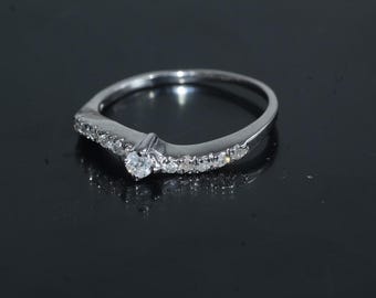 Könnte beinhalten: Ein silberner Ring mit geschwungenem Design, besetzt mit mehreren kleinen, klaren Edelsteinen und einem größeren, zentralen Stein. Der Ring wird auf einer reflektierenden schwarzen Oberfläche präsentiert.