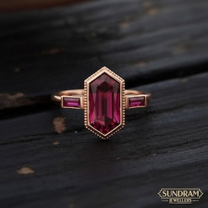 May include: A rose gold ring with a large, rectangular, deep red gemstone at the centre, with two smaller rectangular gems. The central gem is set within a hexagonal frame with a beaded border. The ring is displayed on a dark surface.