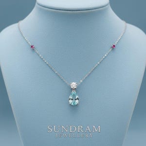 May include: A silver necklace featuring a light blue, pear-shaped gemstone pendant. The necklace has two small red gemstones and a cluster of white gemstones. The text "SUNDRAM JEWELLERY" is visible at the bottom.