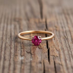 以下が含まれることがあります： 丸い鮮やかな赤い宝石が4本爪でセットされたゴールドリング。素朴な木製の表面に展示されており、シンプルながらエレガントなデザインを際立たせています。バンドは細く磨かれています。