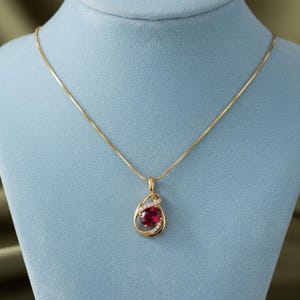 May include: A gold-toned necklace with a teardrop pendant. The pendant features a large, oval-cut, deep red gemstone, encircled by a delicate gold frame and accented with small, sparkling clear stones. The necklace is displayed on a blue mannequin.