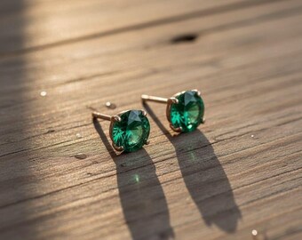 Puede incluir: Un par de pendientes de oro con grandes piedras preciosas redondas de color verde esmeralda. Los pendientes están colocados sobre una superficie de madera, con sombras.