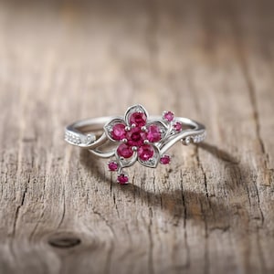 May include: A silver ring featuring a flower design with a cluster of round, bright pink gemstones at the center. Smaller pink gemstones accent the ring's delicate, swirling band. The ring is set against a rustic wooden surface.