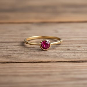 Peut inclure: Une bague en or avec une pierre précieuse rouge vif et ronde au centre. La bague est posée sur une surface en bois, mettant en valeur l'élégance simple du bijou et la couleur vibrante de la pierre. Le jonc de la bague est fin et poli.