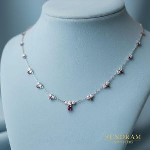 Könnte beinhalten: Eine zarte Silberkette mit Rubin- und Diamantakzenten. Die Kette hat kleine Cluster aus weißen Diamanten und roten Rubinen, mit einem einzelnen Rubinanhänger. Die Kette wird auf einer blauen Schaufensterpuppe präsentiert.